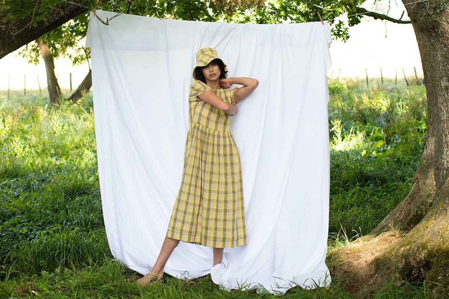 Woman in a yellow gingham dress and bucket hat stands in front of a sheet in a field.